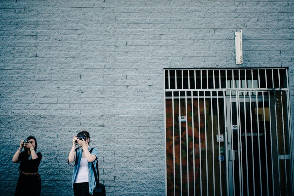 two photography students practicing manual mode in melbourne. they're standing against a light blue wall