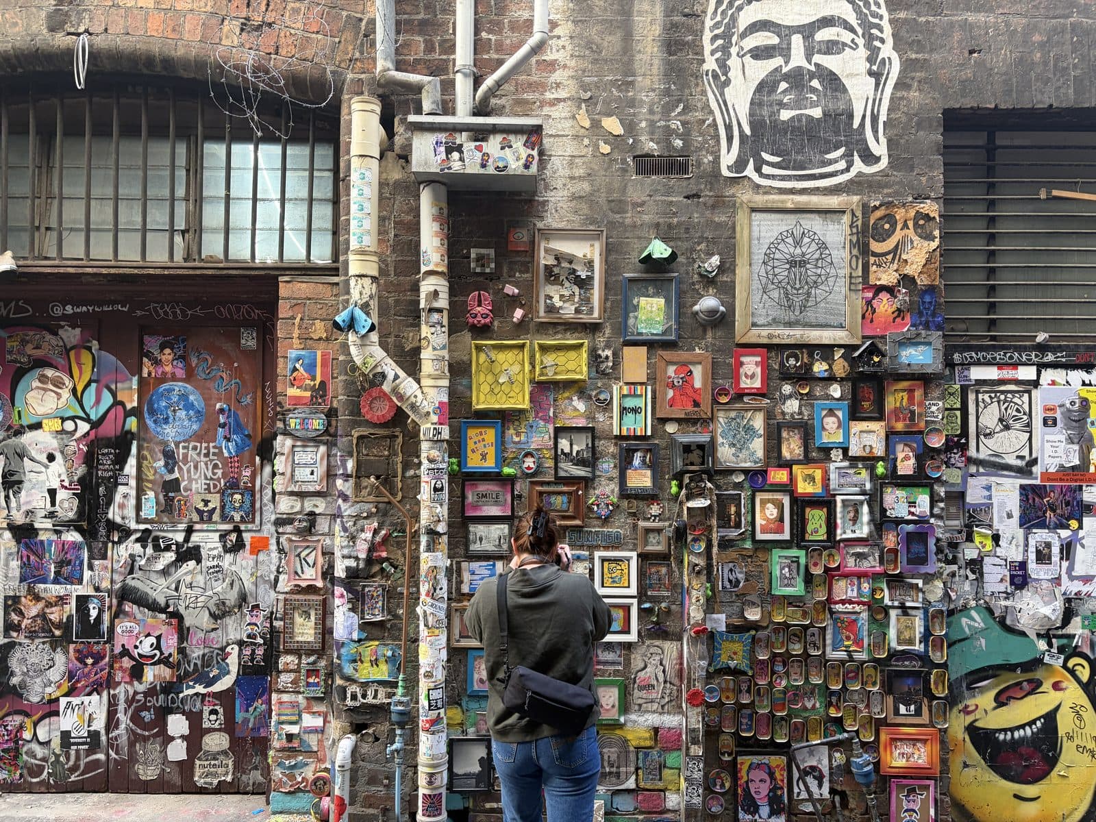 Eliza photographing a brick laneway wall covered in dozens of small framed artworks and stickers in Melbourne CBD