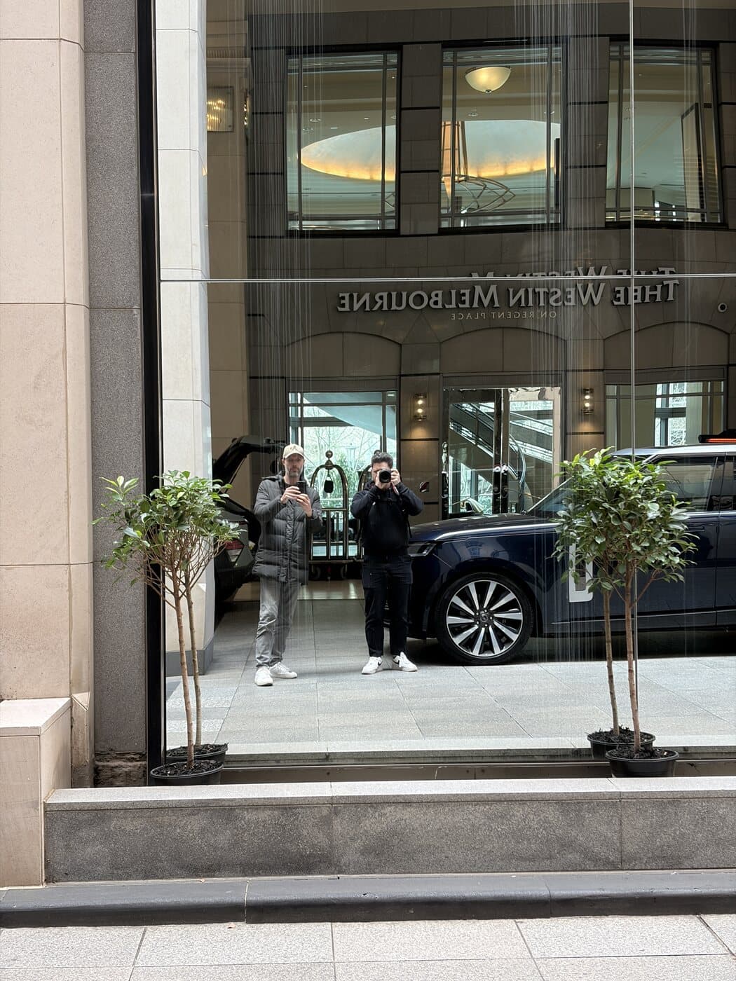 Reflection of two photographers and the Westin Melbourne entrance in a large glass shopfront on Collins Street